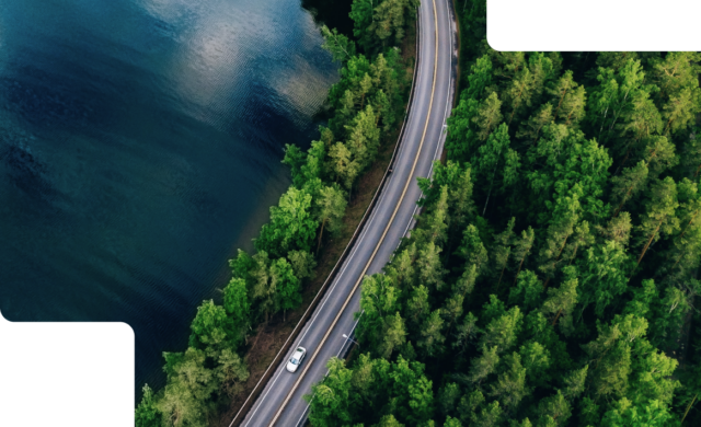 A photograph of a car driving on a long stretch of road with a body of water to the left and a forest to the right.