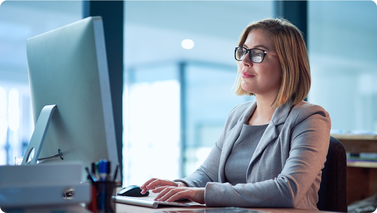 A photograph of a woman sitting at a desk typing on her desktop computer.