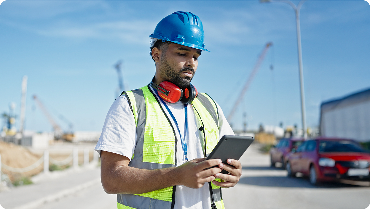 A construction worker wearing a hardhat and safety vest looking at information on a tablet while at an outdoor jobsite.